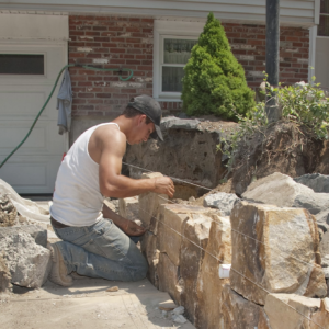 Un albañil con una camisa blanca, jeans y botas de trabajo construyendo una pared de piedra.